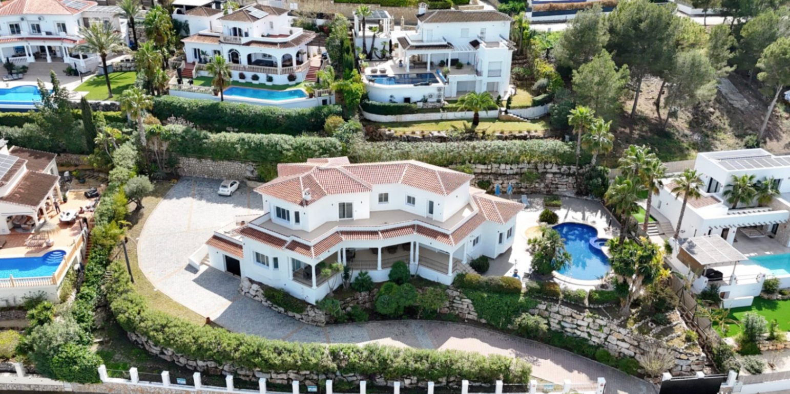 Vista aérea de la villa con fachada blanca y tejado de teja, destacando la amplia zona de aparcamiento pavimentada, muros de piedra y piscina privada rodeada de palmeras.
