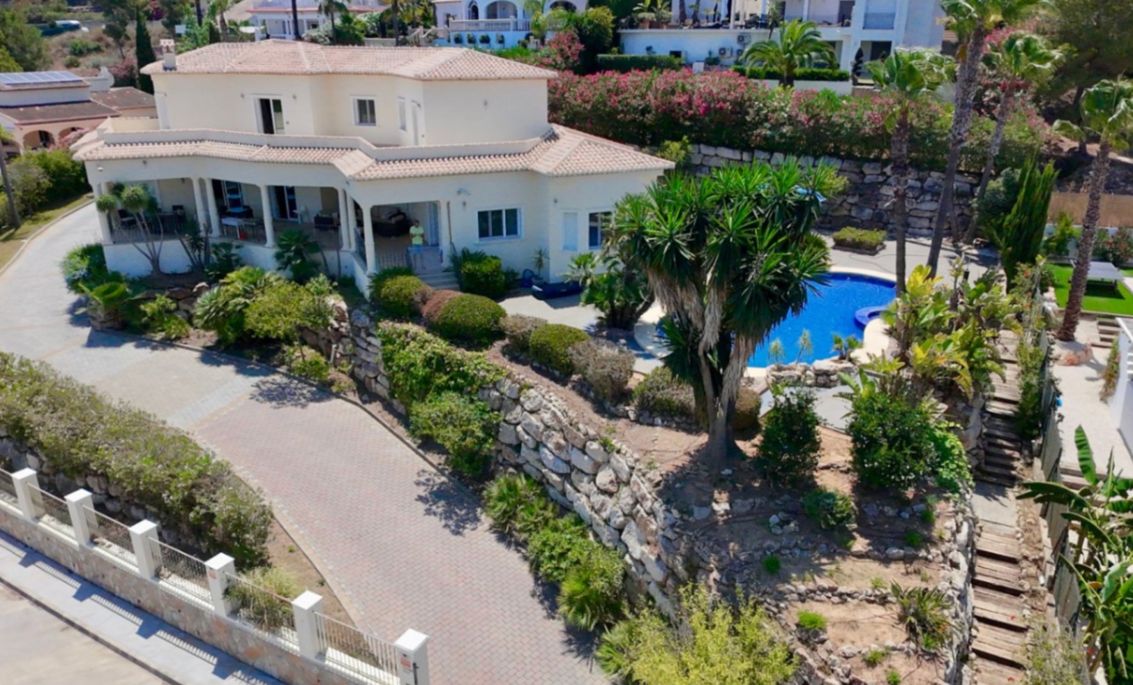 Vista aérea de la villa con fachada mediterránea, amplias terrazas cubiertas, piscina rodeada de palmeras y un jardín aterrazado con muros de piedra natural.