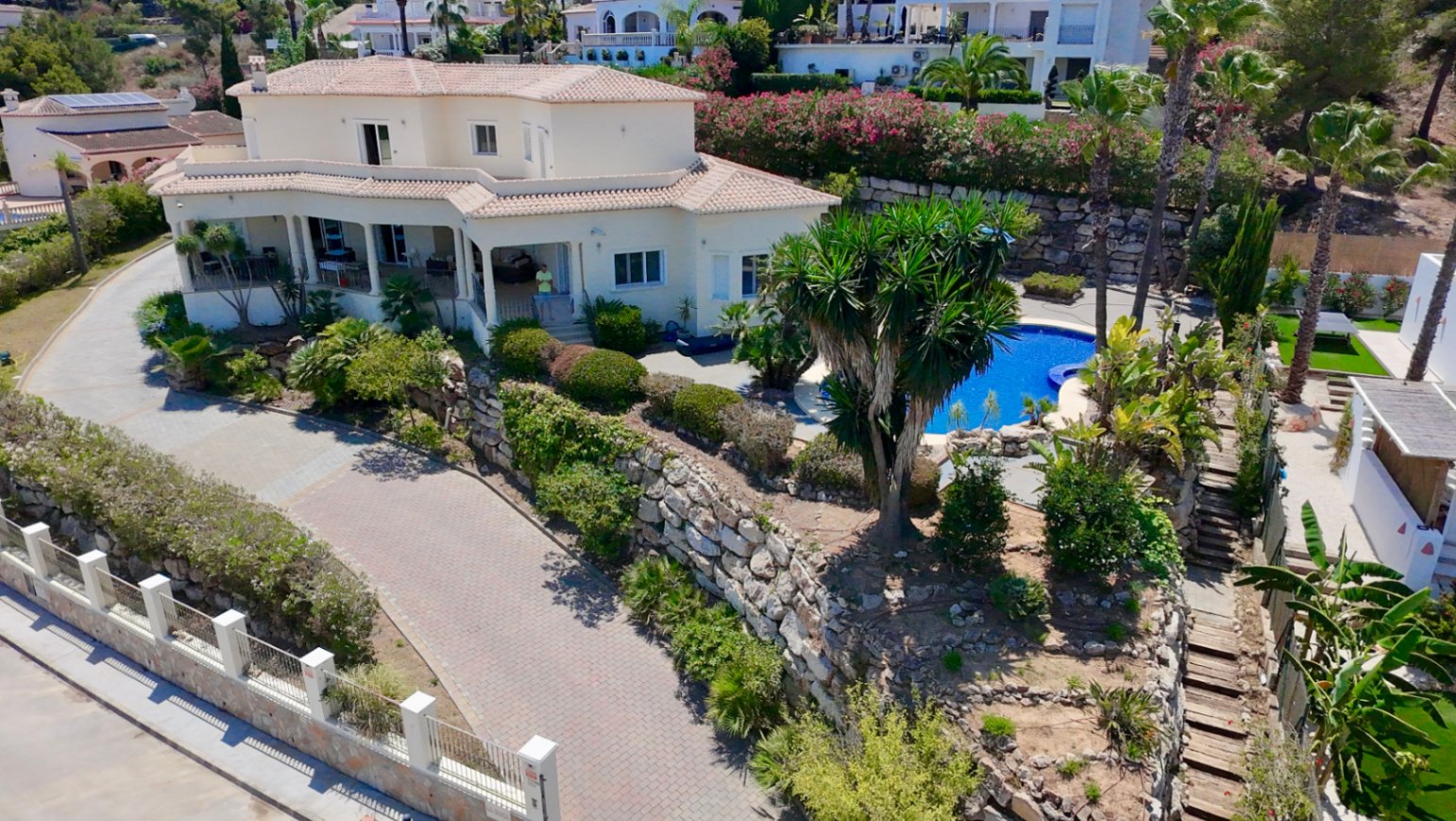 Vista aérea de la villa destacando su arquitectura de dos niveles, la amplia entrada pavimentada y la zona de piscina integrada en un jardín con muros de piedra.