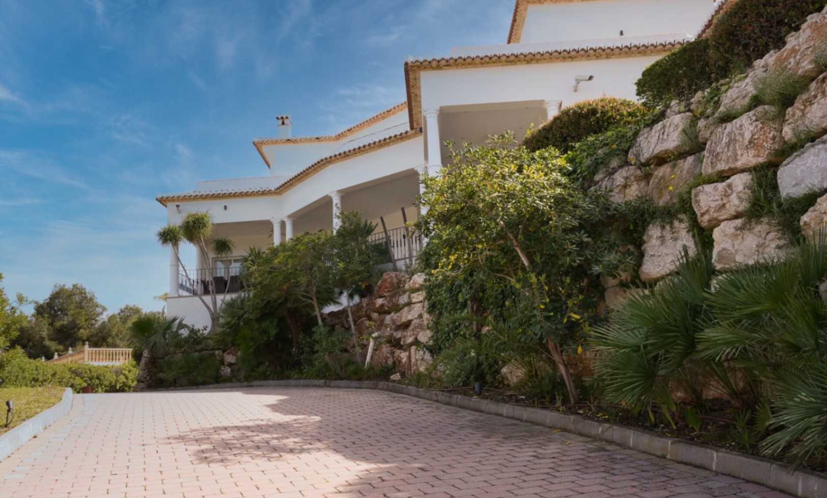 Vista exterior de la fachada blanca con porches de columnas, muros de piedra natural y acceso pavimentado rodeado de vegetación mediterránea.