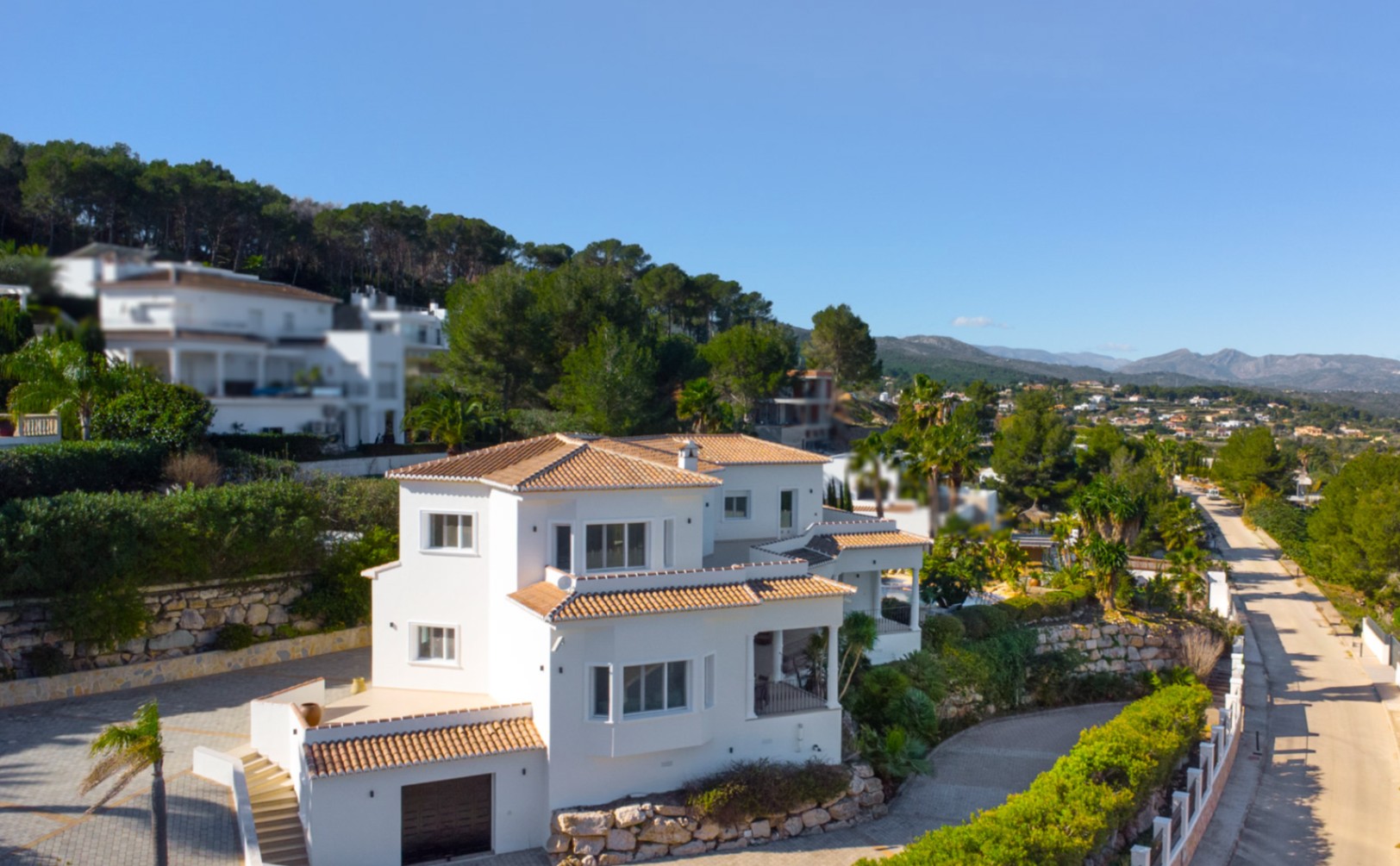 Vista exterior de la villa de estilo mediterráneo con fachada blanca, tejados de teja cerámica y garaje integrado, rodeada de muros de piedra natural en la urbanización Piver.