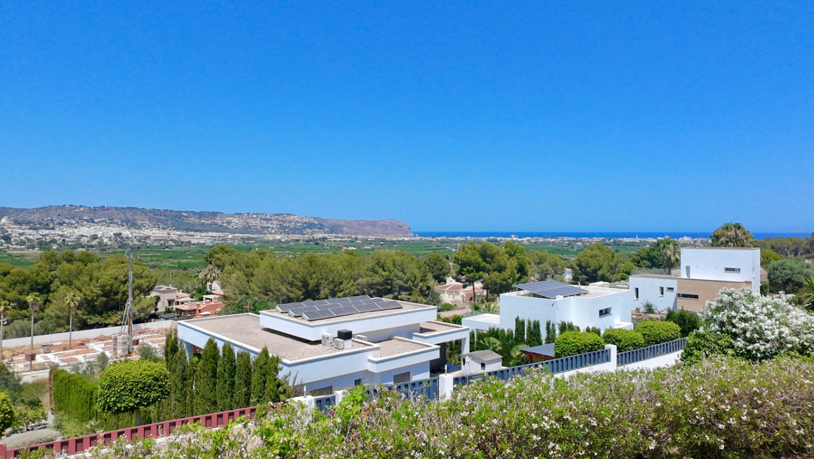 Vista panorámica elevada hacia el mar Mediterráneo y el valle, destacando la arquitectura moderna de las villas circundantes con paneles solares y vegetación autóctona.